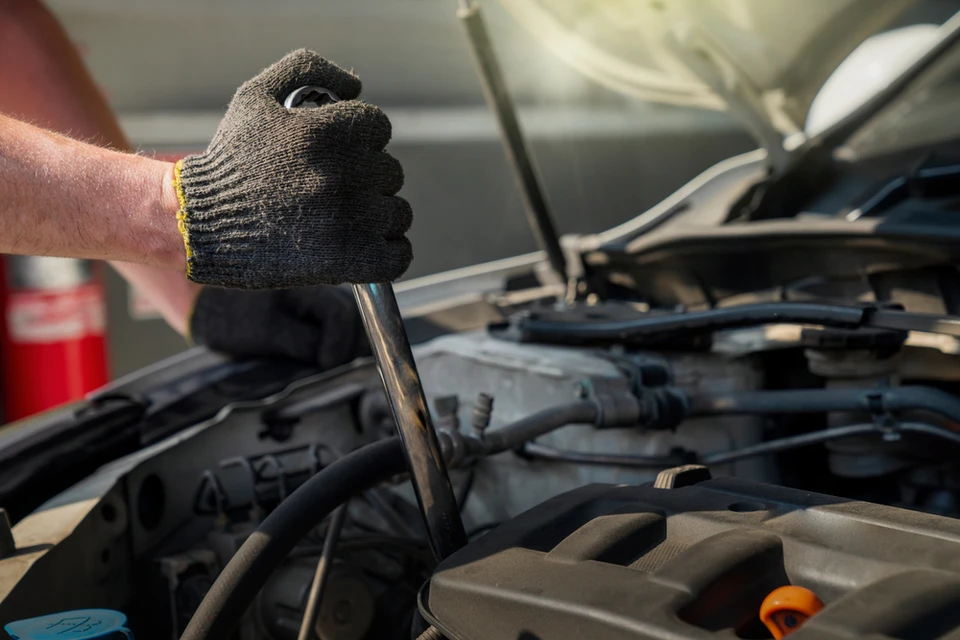 Photographie en couleur montrant l'intérieur d'un atelier automobile moderne et bien équipé. Au premier plan, un mécanicien qualifié en combinaison de travail bleu marine, portant des gants de protection noirs, inspecte ou travaille sur le moteur d'une voiture surélevée sur un pont élévateur. L'atelier en arrière-plan est propre et organisé, avec des outils rangés méthodiquement sur les murs, un éclairage LED professionnel et du matériel de diagnostic visible. L'atmosphère doit transmettre la compétence, la confiance et la transparence du travail professionnel en mécanique automobile. Pas de personnes souriantes de manière artificielle, uniquement un professionnel concentré sur son travail.
