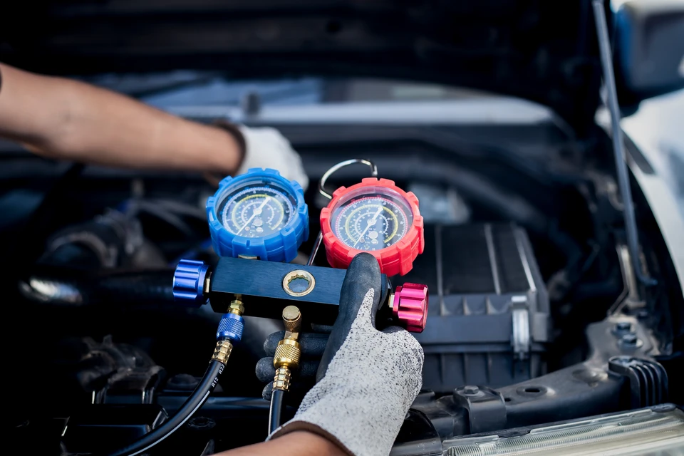 Mécanicien automobile en tenue professionnelle travaillant sur un système de climatisation automobile à l'intérieur d'un atelier moderne bien équipé. Le professionnel manipule des connectiques de recharge de climatisation (manomètres, flexible de recharge) sur un véhicule en panne ouverte ou en soubassement visible. Environnement d'atelier propre et professionnel avec outillage et équipements de diagnostic climatisation en arrière-plan. Lumière naturelle ou d'atelier professionnel. Accent sur le professionnalisme et la technicité du service de climatisation automobile.